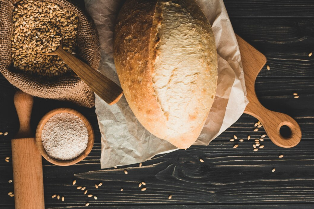 Freshly baked bread loaf on a wooden board with flour and ingredients in the background.